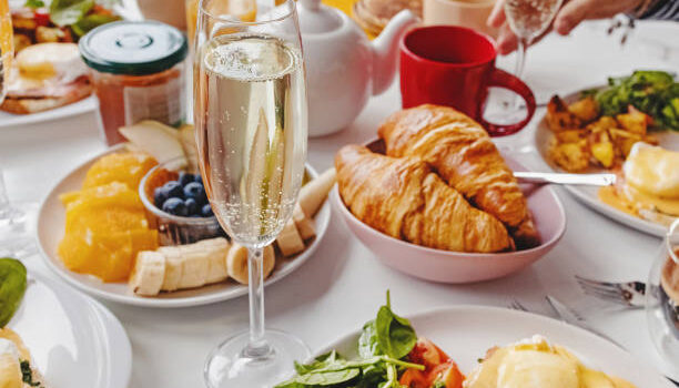 Glass of champagne close-up on the table with different breakfast and brunch dishes like croissants, eggs benedict, coffee and fruits. Selective focus