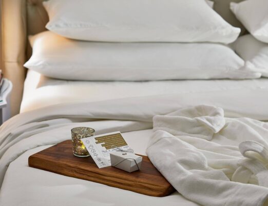 a smooth wood board sitting on a hotel room bed with a note, a gift box, and a candle on top
