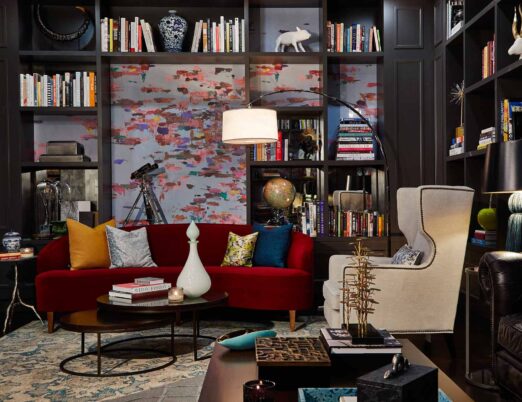 An eclectic sitting room in a luxury hotel with an elegant red couch and beige chair and many shelves of books