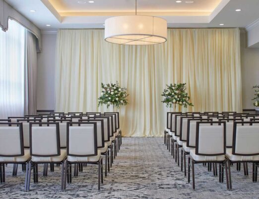 conference room with rows of brown and white chairs with curtains on each wall and two flower arrangements at the front of the room