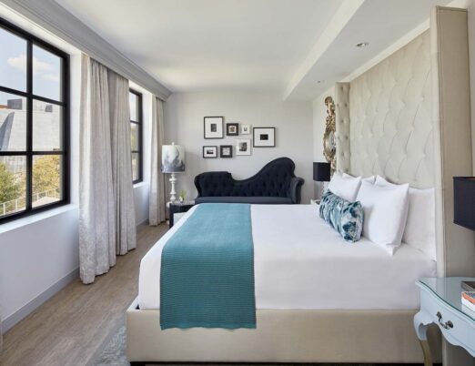 A long luxury hotel room with king bed with white linens, two large windows, and a plush day bed