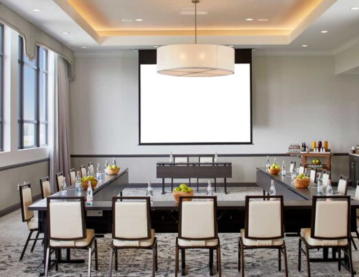 boardoom with L shaped covered with fruits and water bottles table surrounded by brown and cream chairs, facing a blank projector screen