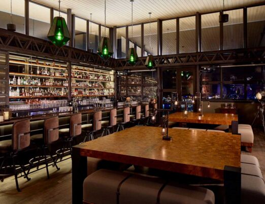 an indoor roof top bar with dozens of bottles, and green pendent lights surrounded by floor-to-ceiling windows