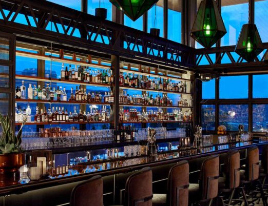 an indoor roof top bar with dozens of bottles, and green pendent lights surrounded by floor-to-ceiling windows