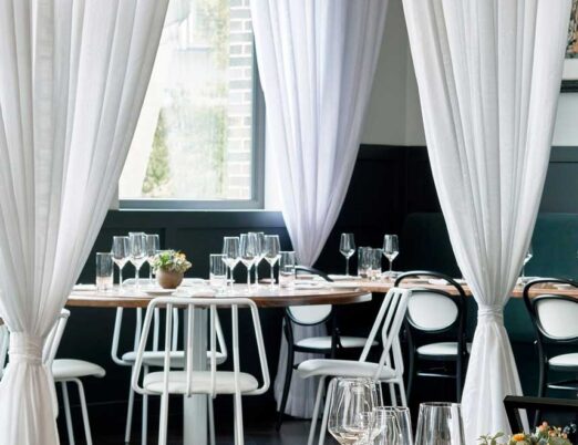 bright restaurant with white curtains, white chairs, and wooden tables with glasses and flowers on them