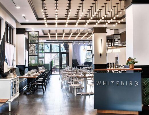 bright restaurant with white curtains, white chairs, and wooden tables with glasses and flowers on them