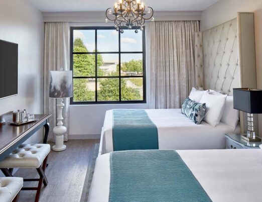 A luxury hotel room with two double beds, chandelier, flat screen tv, and a large window