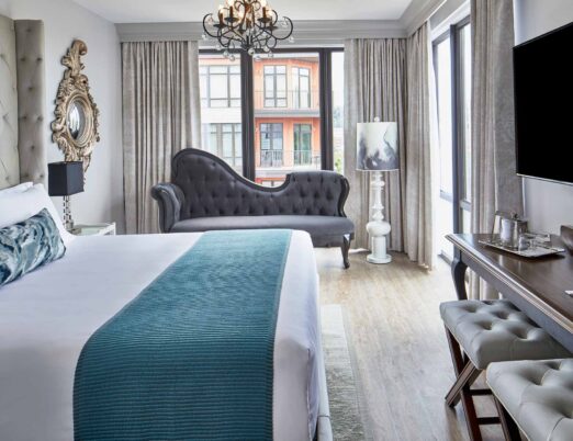 a bright luxury hotel room with a king-size bed with white linens, a chandelier, a plush day bed, and a flat screen tv