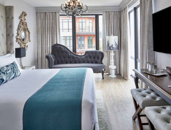 a bright luxury hotel room with a king-size bed with white linens, a chandelier, a plush day bed, and a flat screen tv