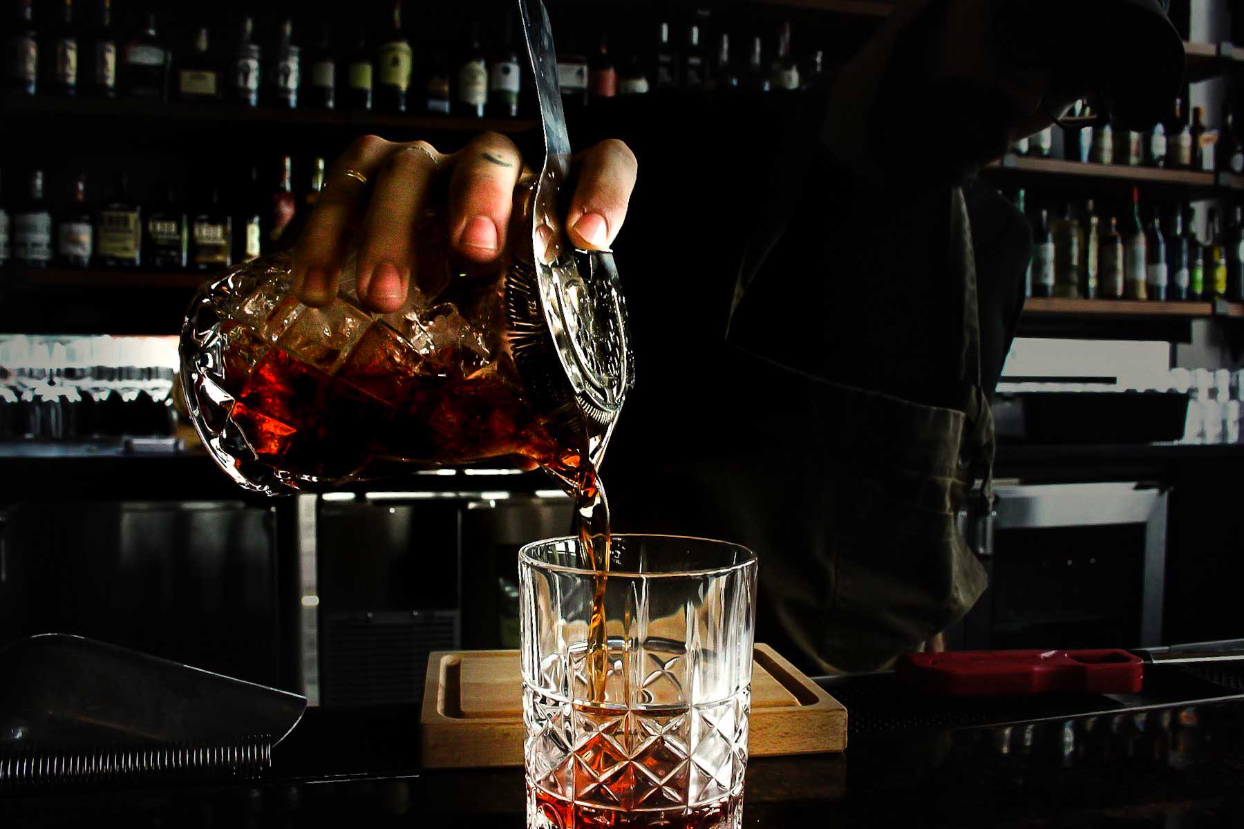 a bartender pouring a cocktail through a strainer into a crystal glass