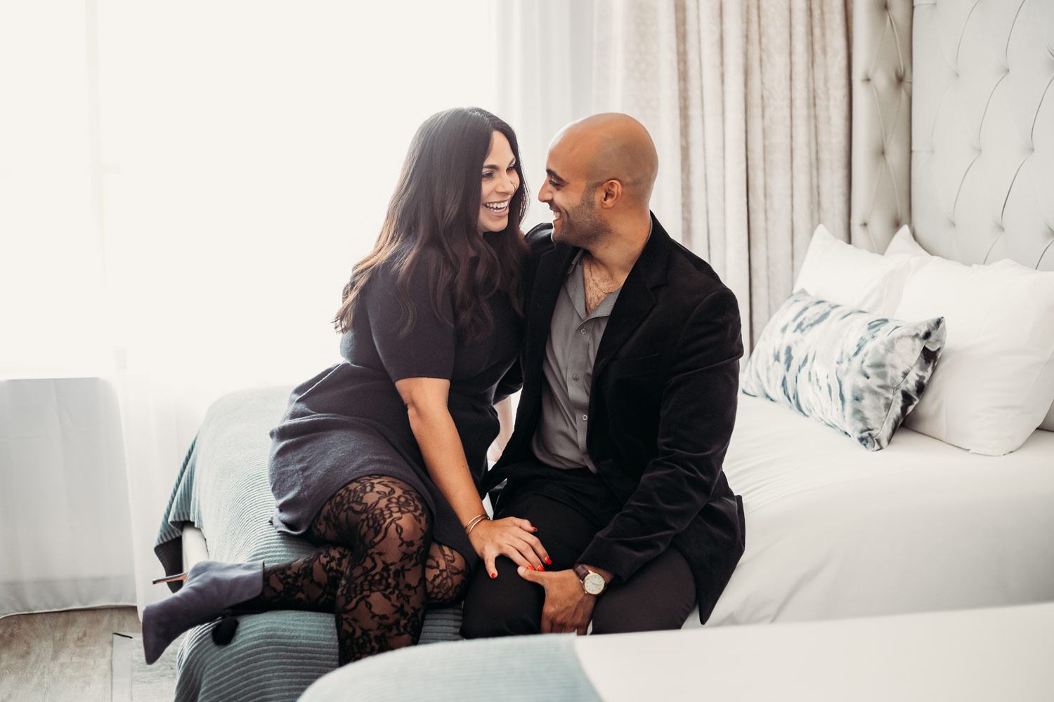 couple smiling while sitting on hotel bed with one another