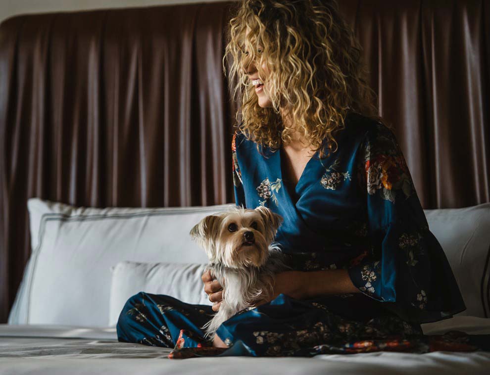 dog and woman with dirty blonde curly hair sitting on bed with white sheets and pillows