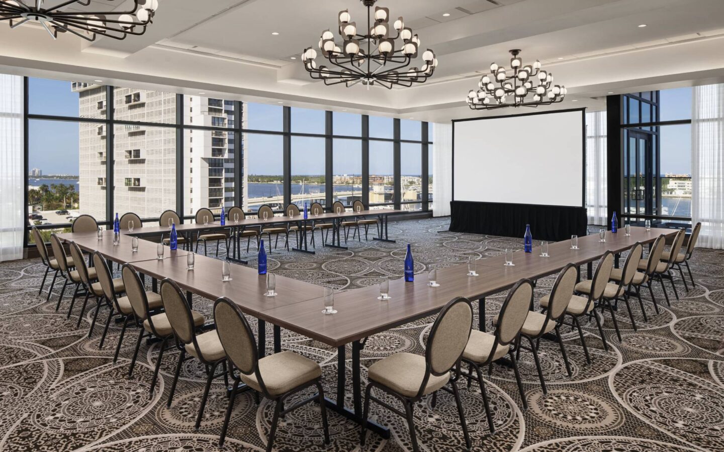 Conference room set with U-shaped tables, chairs, water bottles, a projection screen, chandeliers, and large windows overlooking the city.