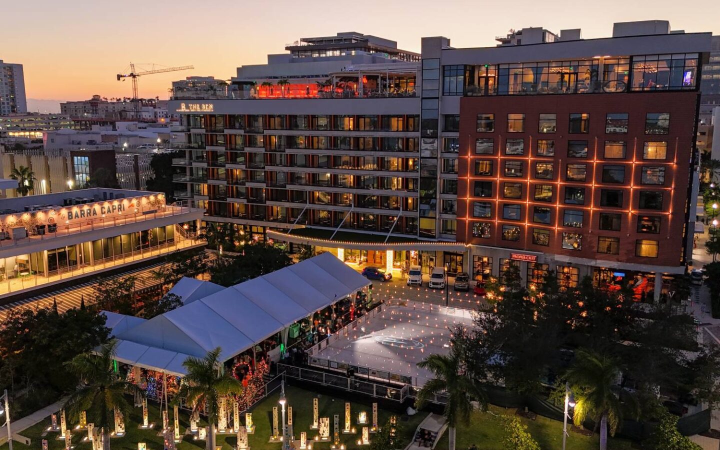Aerial view of an outdoor ice skating rink and holiday market surrounded by illuminated buildings at sunset.