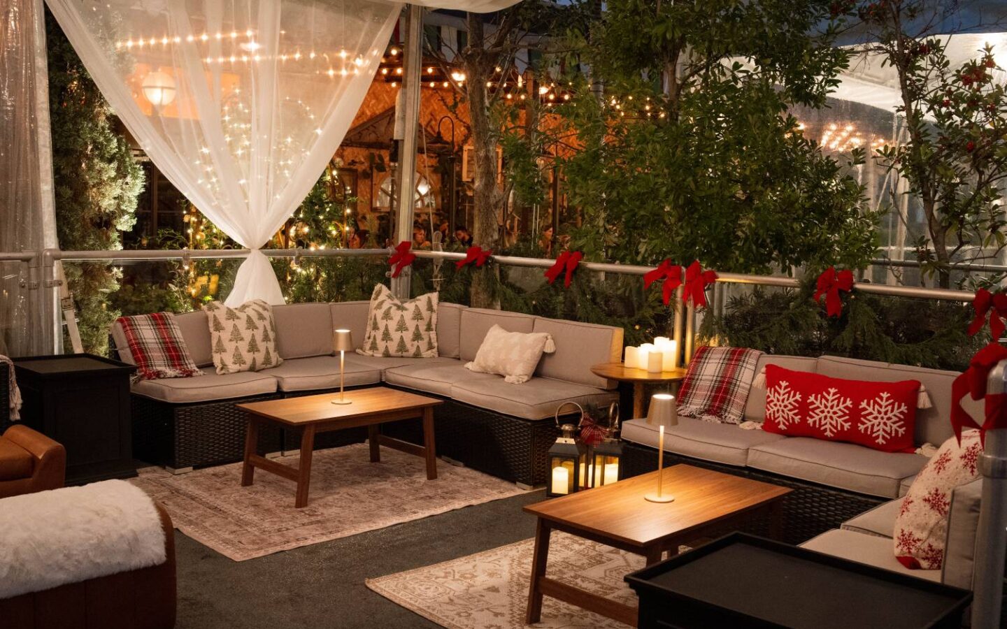 Cozy holiday lounge with festive pillows, red bows, candles and string lights surrounded by greenery.