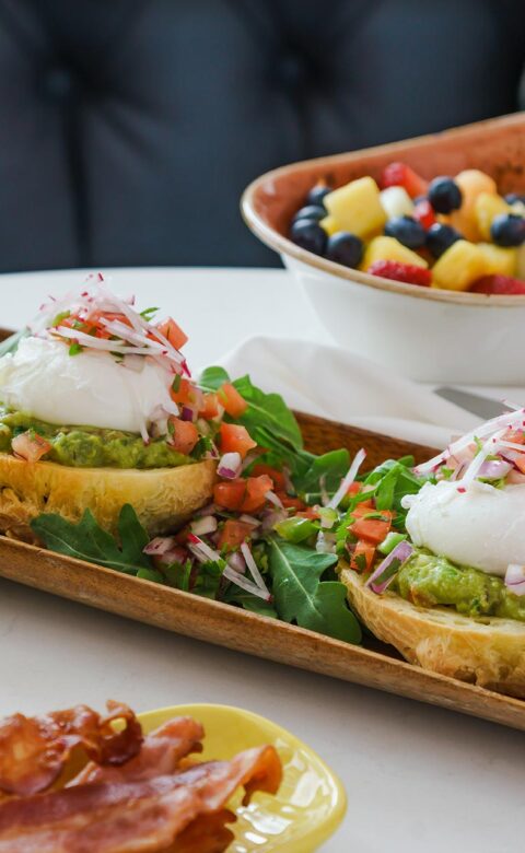 eggs benedict with avocado topped with diced tomatoes and onions beside a bowl a fruit and a side of bacon