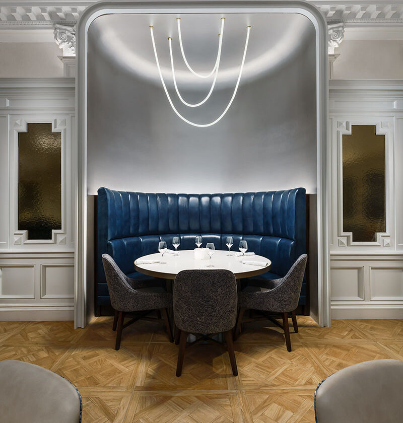 a round dining table set into an enclave in the wall of a hotel dining room with 3 chairs and high-backed blue leather booth seat