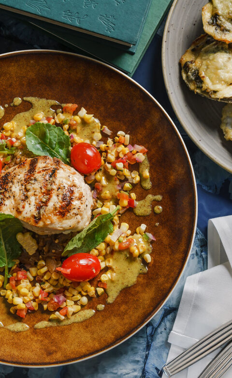 a fine dining dish of swordfish, corn, and cherry tomatoes beside a plate of baked oysters