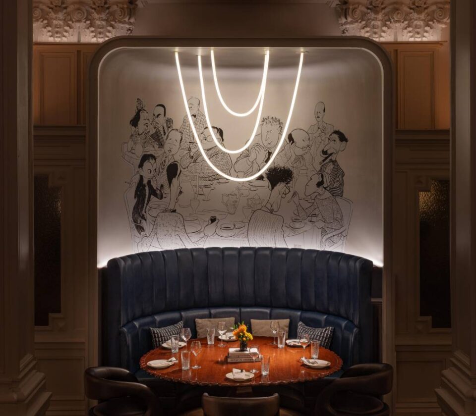 Elegant restaurant dining alcove with curved blue banquette seating, a set table, statement lighting and illustrated wall art.