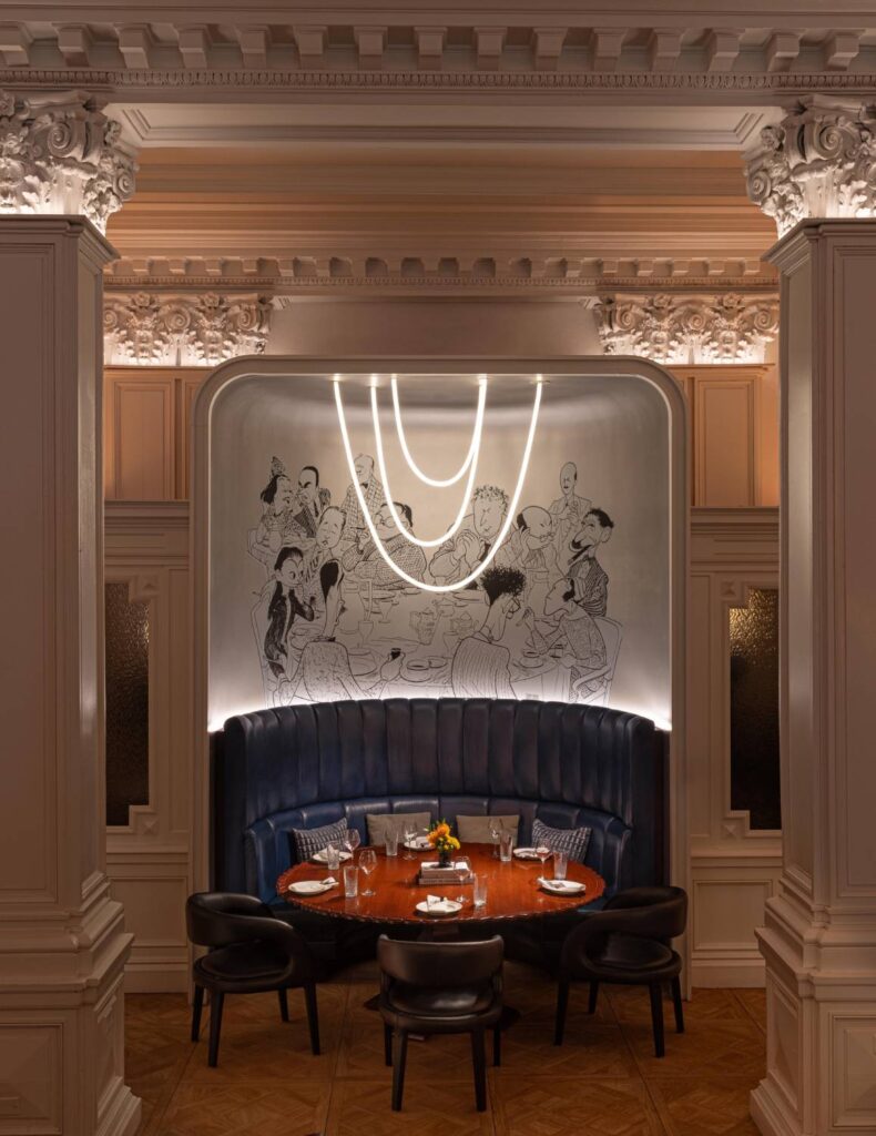 Elegant restaurant dining alcove with curved blue banquette seating, a set table, statement lighting and illustrated wall art.
