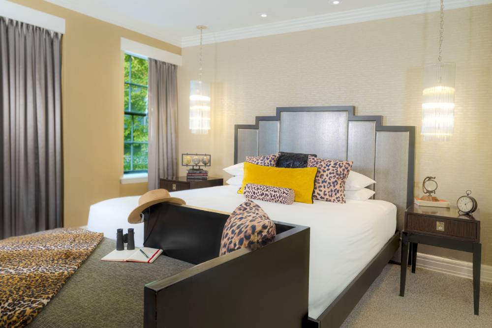 Hotel bedroom with a king bed, leopard-print accents, yellow pillows, pendant lights, and grey curtains.