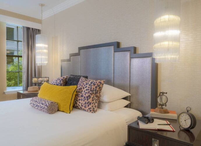 Hotel bed with leopard-print and yellow accent pillows, pendant lights, and a bedside table with vintage-style decor.
