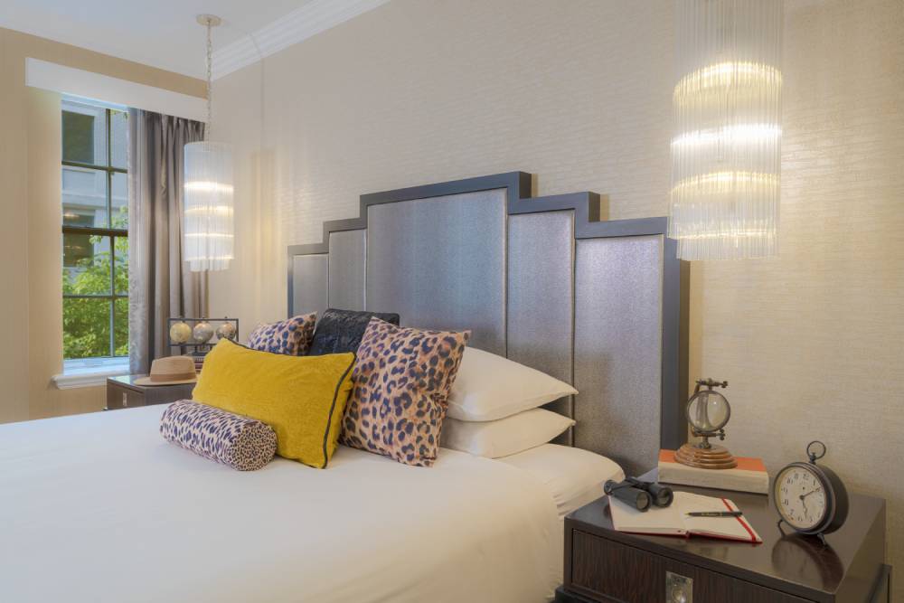 Hotel bed with leopard-print and yellow accent pillows, pendant lights, and a bedside table with vintage-style decor.