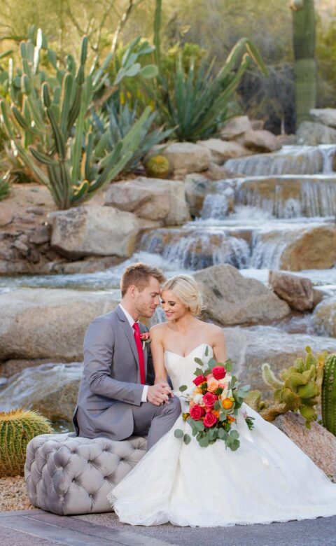 bride and groom sit on rocks next to cactus lined waterfall