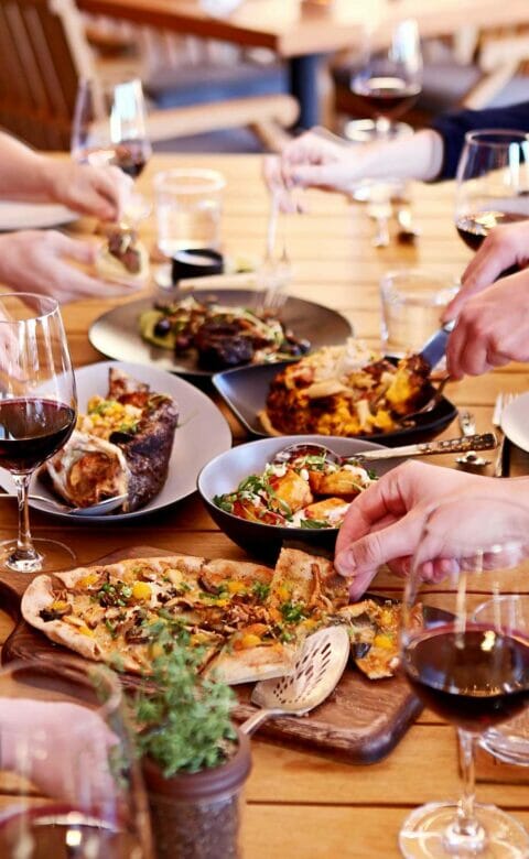 wooden dining table covered with plates of food and wine glasses being used by people