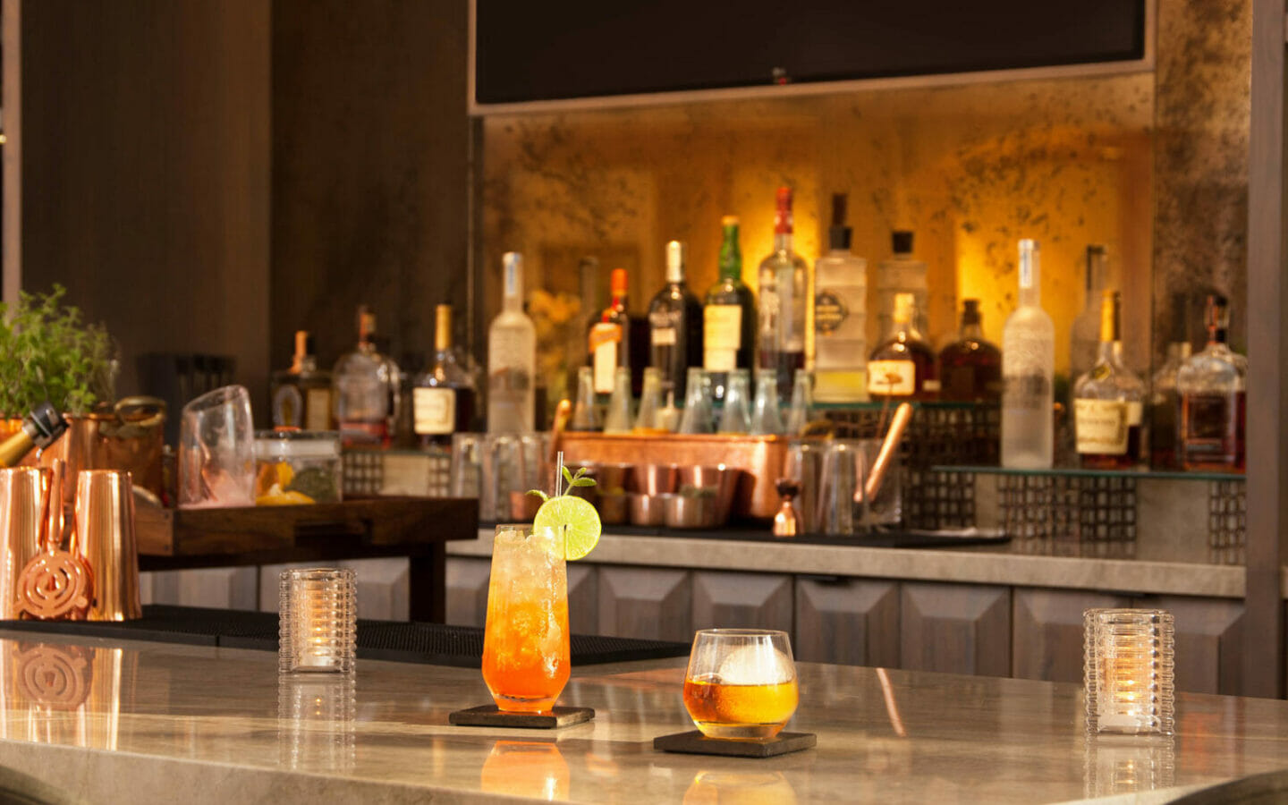bar counter with drink in tall glass and drink in small glass with various bottles on the shelf in the background