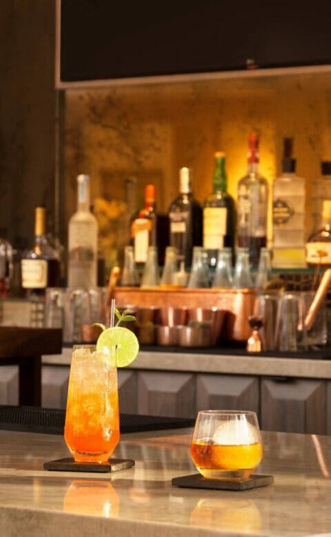 bar counter with drink in tall glass and drink in small glass with various bottles on the shelf in the background