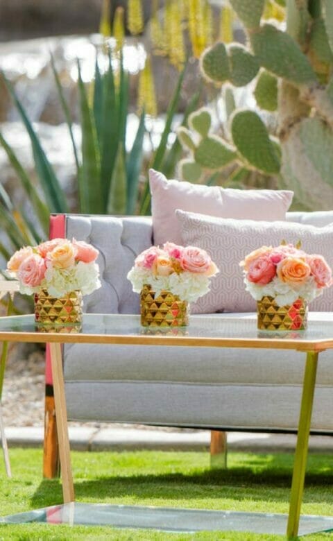 sitting area at outdoor wedding venue with couch and two arm chairs. glass gold coffee table is decorated with three small bouquets of pastel roses next to large cacti