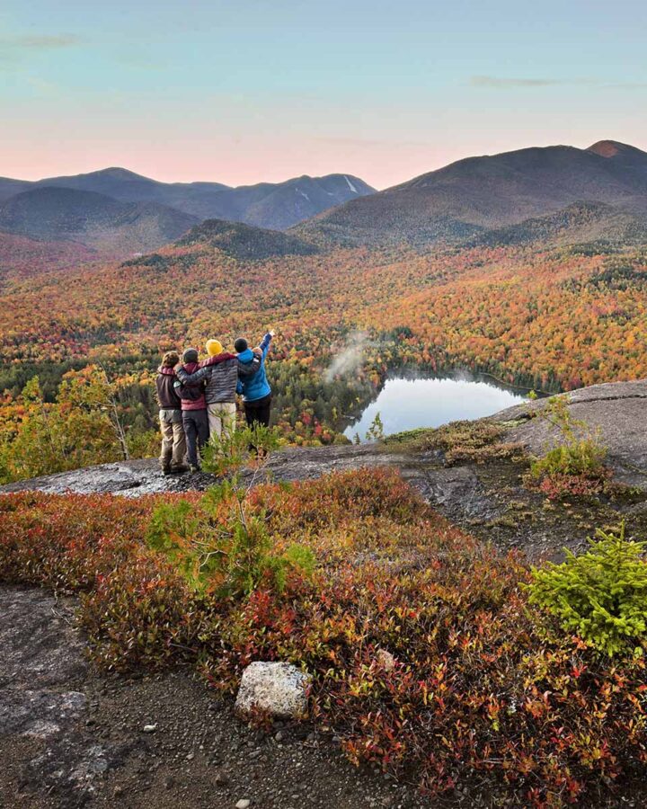 group of four friends stand on mountain peak during fall and pointing to view